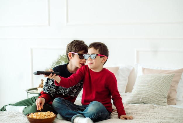 Симпатичные дети едят попкорн, смотря телевизор дома в трехмерных очках.