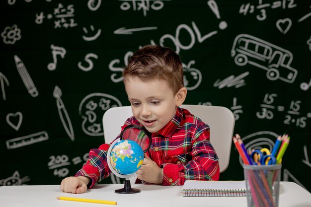 Cute clever boy is sitting at a desk with magnifying glass in hand. Child is reading a book with a blackboard on a background. Ready for school. Back to school