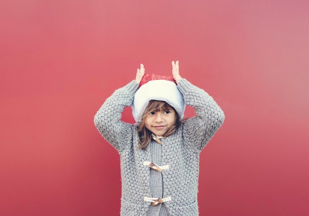 Cute confident little girl wearing santa claus hat putting hands like the reindeer horns Funny kid playing against red wall outdoors in Christmas time Happy xmas and New Year 2021 Xmas holiday