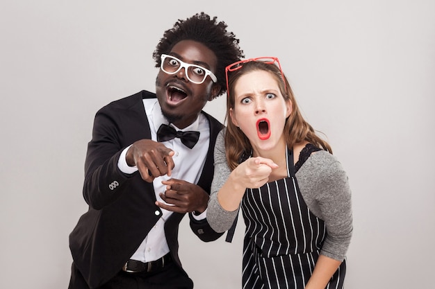 Cute couple pointing fingers at camera and have a shocked look. Studio shot, gray background