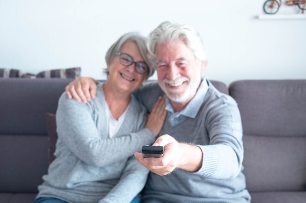 Cute couple of two caucasia mature seniors o the sofa watching  a film or something in the TV - man holding the remote control