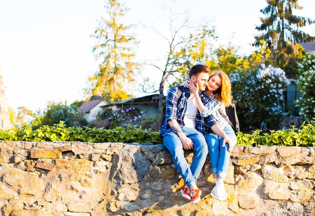 The cute couple with backpacks on their shoulders, sitting on an old stonewall during the sunset