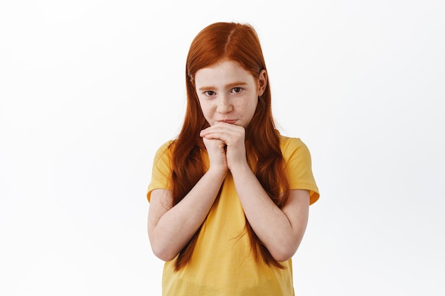 Cute coy redhead girl with freckles begging for something, say please with adorable face expression, being needy, standing over white background