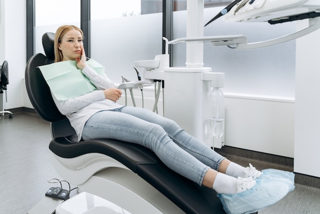 Cute, crooked woman touches sitting in a dental chair. The woman touches her cheek, she has a toothache