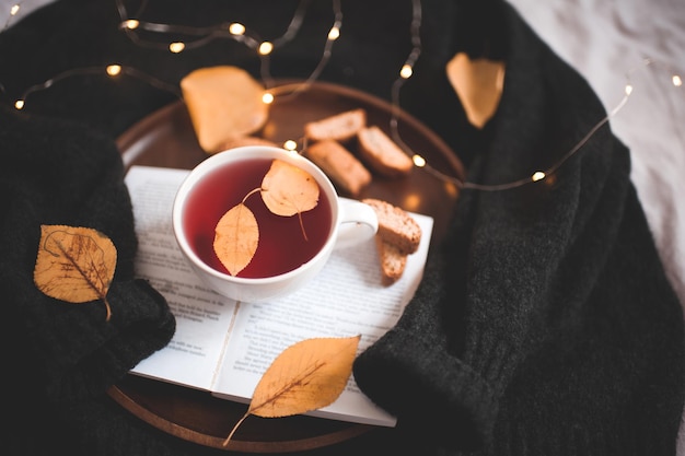 Cute cup of tea with fallen leaves on open book closeup over glowing lights. Good morning. Breakfast.