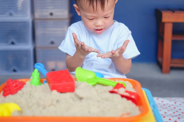 Милый любознательный азиатский малыш 2-х лет, играющий с кинетическим песком в песочнице дома / в детской / дневной уход