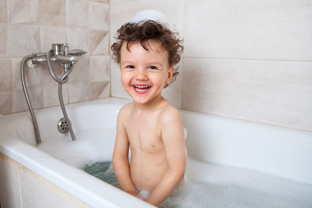 Cute curly baby bathes in the bath with an emotional laugh. Personal hygiene rules for children. Boy, a child plays with toys in a full bath with water.