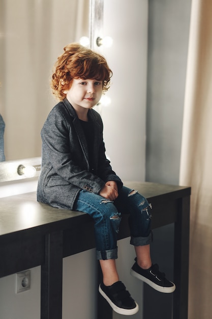 Photo cute curly boy child in jacket and ripped jeans posing near the mirror and smiling