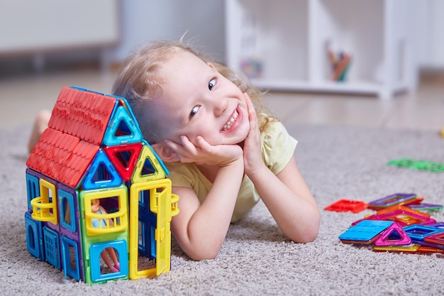 写真 子供部屋のカーペットの上に自宅の磁気コンストラクターで作られた家の横にあるかわいい巻き毛の女の子