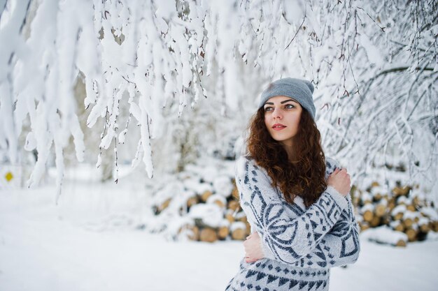 Carina ragazza riccia in maglione e copricapo al parco forestale innevato in inverno.