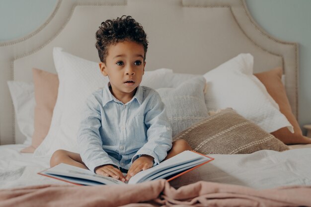 Cute curly haired afro american little boy sitting on bed with book