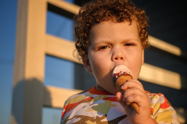 Cute curly haired boy eating ice cream waffle cone outside on a summer Sunny day