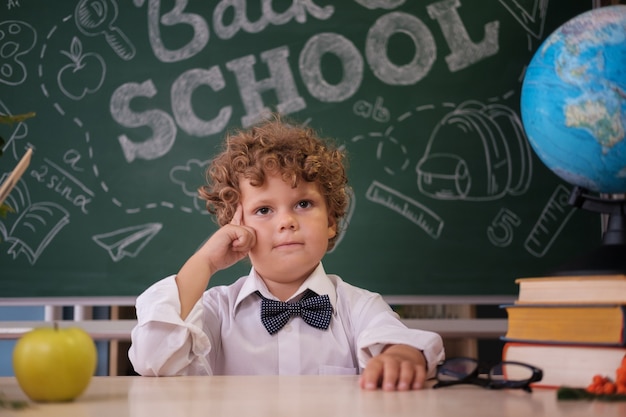 A cute curly-haired boy is sitting at a desk against the background of a blackboard with the inscription in English back to school