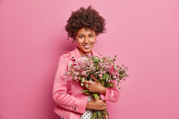 Cute curly haired woman embraces big bouquet of flowers recieved from boyfriend has festive mood wears jacket isolated over pink wall