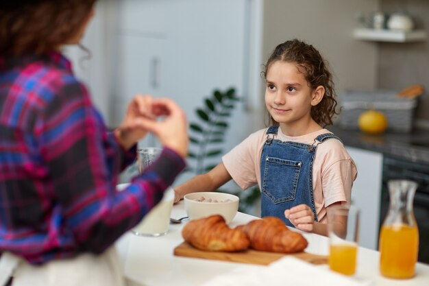 手でハートのシンボルの形をしている笑顔のお母さんを見て、キッチンでかわいい巻き毛の少女。