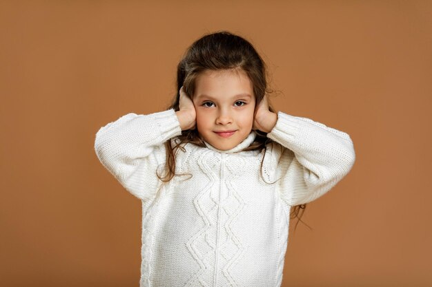 Милая кудрявая маленькая девочка в белом свитере закрывает уши