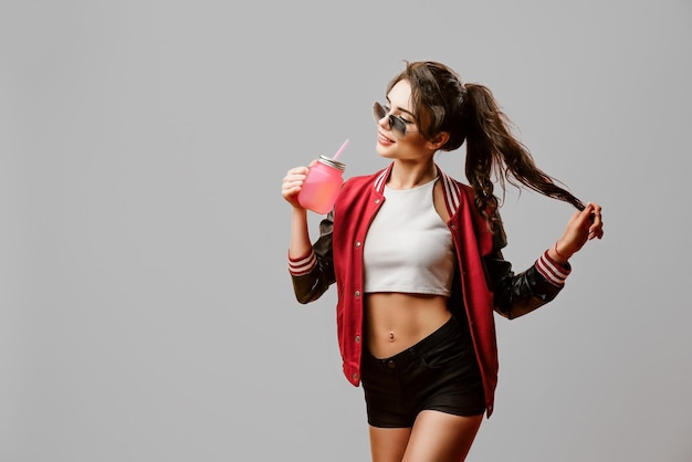 Cute curly stylish young woman in sunglasses holds beverage in glass over gray background