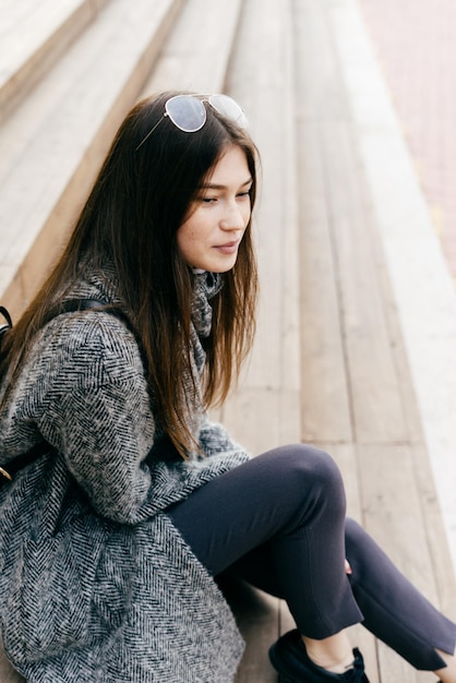 Cute dark-haired girl in gray coat sits on steps in the open air, street style and fashion