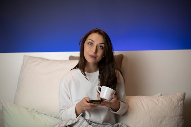 Cute dark-haired girl sitting in bed with phone cup of coffee looking at camera