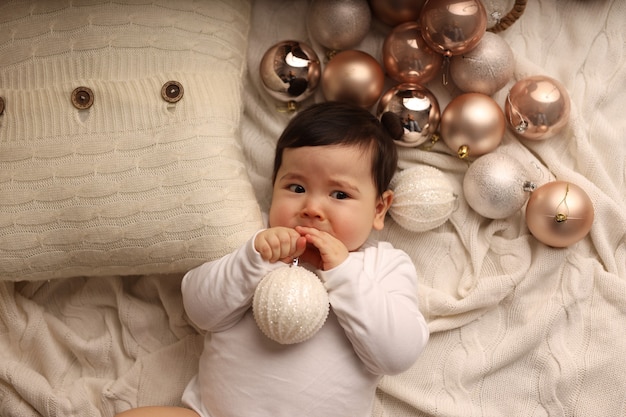 A cute darkskinned girl lies on a white knitted blanket and plays with Christmas tree toys