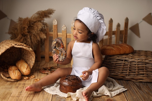 A cute darkskinned kid with curly hair in a chefs costume eats a bagel and a roll