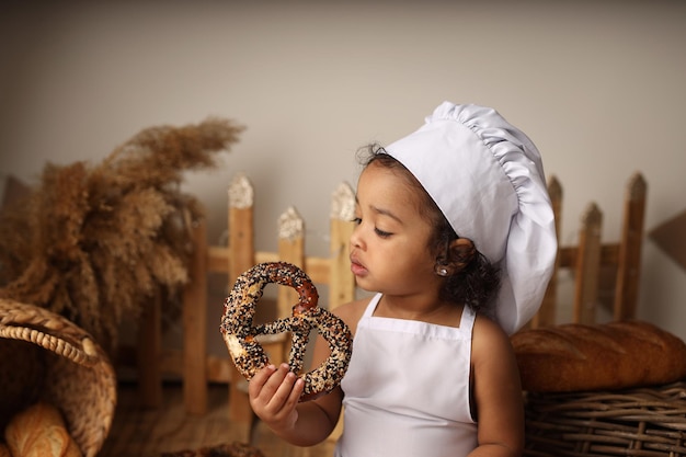 A cute darkskinned kid with curly hair in a chefs costume eats a bagel and a roll