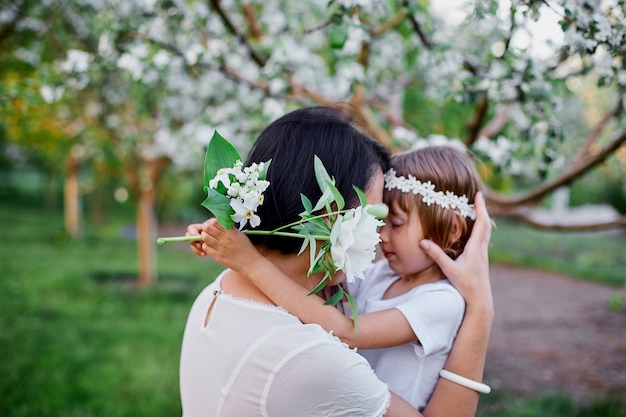 花の春の庭で抱き締めるかわいい娘と母幸せな女性と子供、屋外で白いドレスを着て、春の季節が来ています。母の日の休日の概念