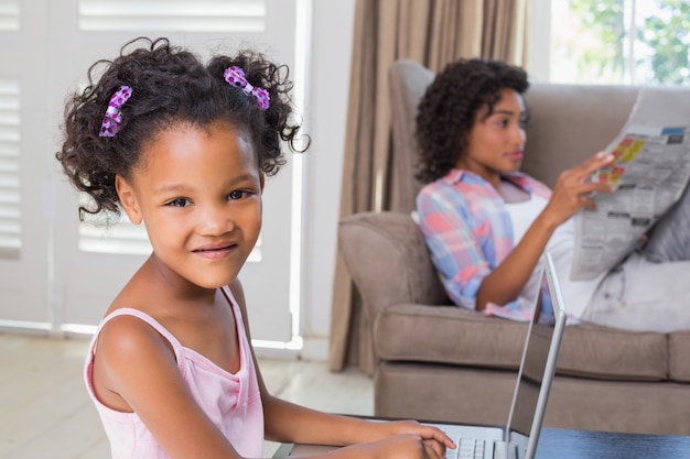 Cute daughter using laptop at desk with mother on couch