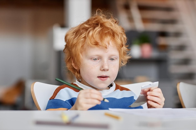 Cute diligent gingerhaired learner of nursery school with\
crayon of dark green color drawing by desk