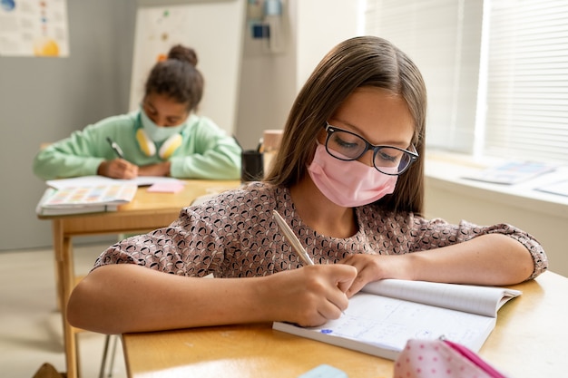 Studentessa diligente carina in maschera protettiva che prende appunti in quaderno a lezione