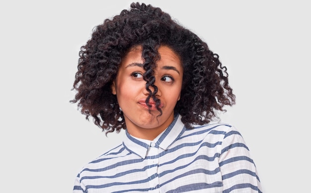 Cute doubtful African American young woman looking aside with dissatisfied face dressed in casual striped shirt posing over white wall Student girl has confused expression because of hairstyle