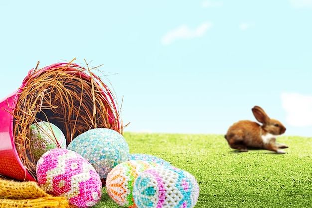 Cute easter bunny and colorful easter eggs spilled from the nest in a red bucket with fabric on the field. Happy Easter