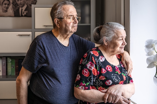 A cute elderly couple stands at the window and looks out for someone, waiting.