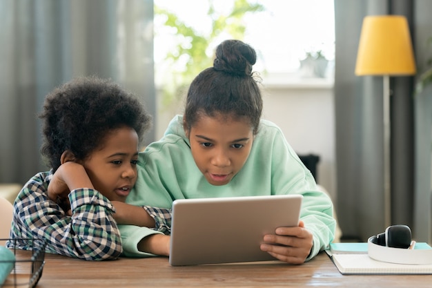 Cute elementary mixed-race siblings sitting by table in living-room and watching online educational video while preparing homework