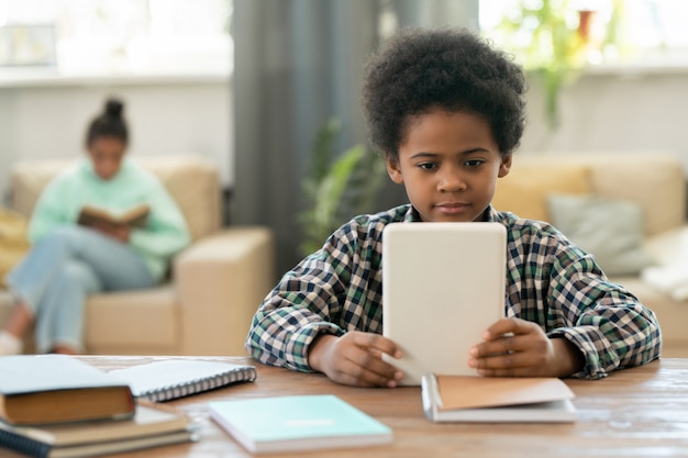 Cute elementary schoolboy of African ethnicity sitting by table in living-room against his sister and watching online educational video