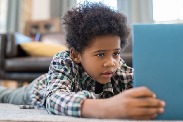 Cute elementary schoolboy with digital tablet lying on the floor against black leather couch in living-room and watching curious online video