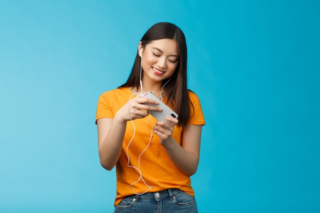 Carino intrattenuto bella studentessa urbana trascorrere la pausa giocando a giochi di corse, tenere smartphone inclinazione orizzontale telefono spostare auto, sorridente ampiamente in piedi entusiasta sfondo blu.