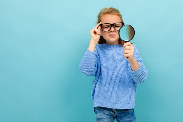 Cute european girl in glasses looks through a magnifier on light blue