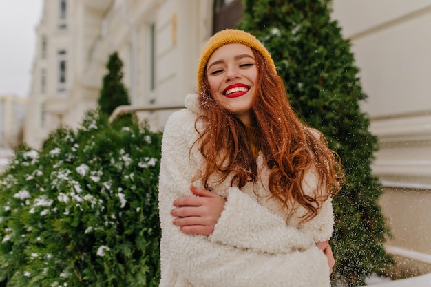 Cute european girl in trendy coat enjoying winter Lovely redhaired woman posing with sincere smile in cold day