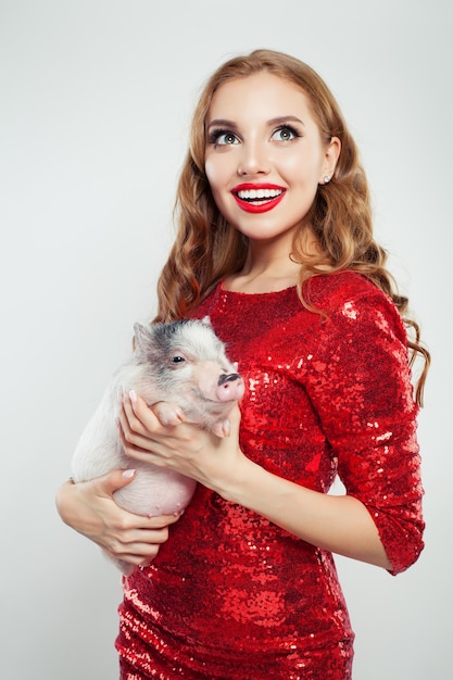 Cute excited woman in red fashionable dress holding mini pig on white background