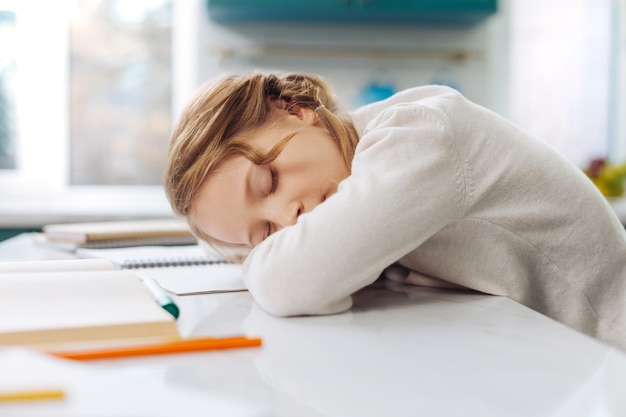 Cute fair-haired little schoolgirl sleeping over her books while sitting at the table and doing homework