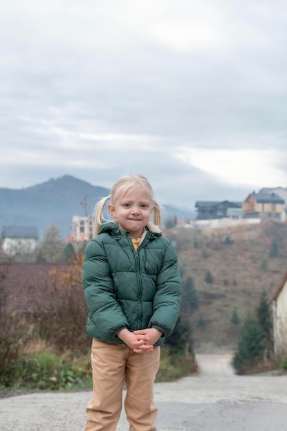 Cute fairhaired girl with two ponytails wears warm green autumn jacket mountain background Vertical frame