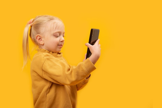 Cute fairhaired kid with two ponytails girl takes selfies preschooler in yellow jacket looks at phone and smiles