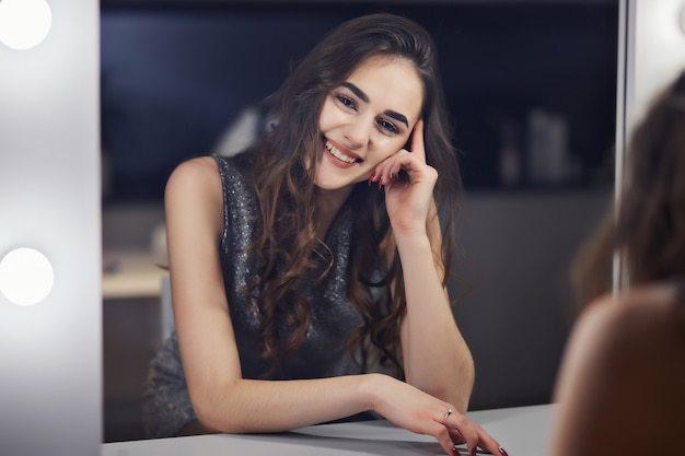 Cute fashion woman with long brunette curly hair looks in salon mirror.