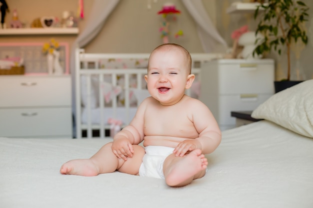 Cute fat toddler girl smiles in diapers sitting on bed at home