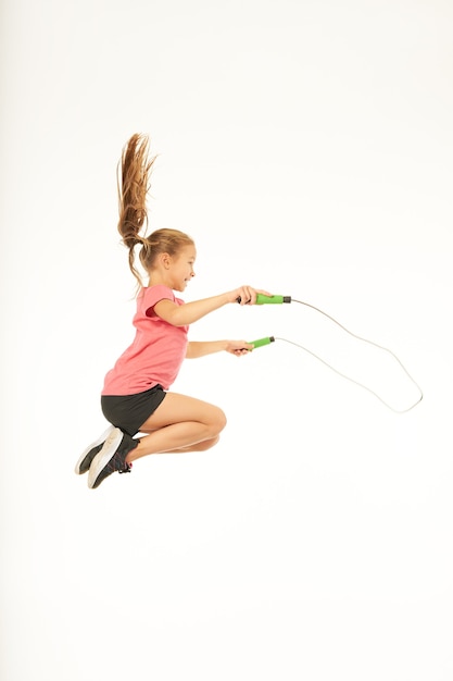 Фото Милый ребенок женского пола в спортивной одежде смотрит в сторону и улыбается, прыгая через скакалку в воздухе. изолированные на белом фоне