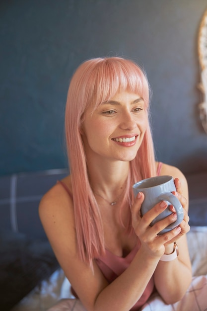 Cute female model holds cup of delicious drink sitting on bed