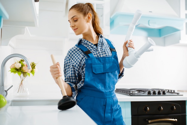 制服を着たかわいい女性配管工が台所でプランジャーとパイプ、詰まりを保持しています。ハンディウーマン修理シンク、家庭用衛生設備サービス