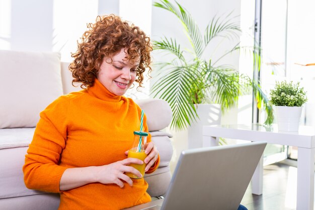 Cute female red hair freelancer reading notebook while sitting on floor with open computer in modern home, young creative woman work on laptop while having orange juice