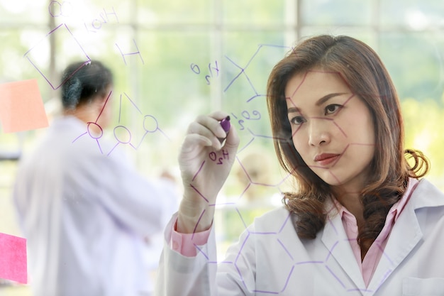 Cute female scientist with marker writing chemical formulas on glass board while working with colleagues in laboratory.